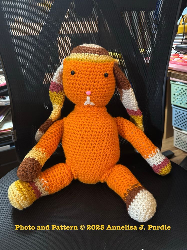An orange crocheted bunny with long ears and yellow accents.  He sits up in a chair facing the viewer.
