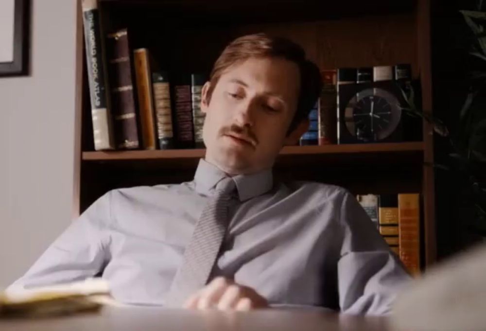 a man sits at a desk in front of a bookshelf with a world book atlas on it