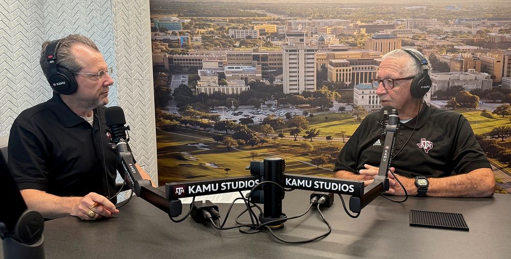 KAMU host Jay Socol and guest Dave South, seated, wearing headphones and speaking into microphones, record a discussion inside the KAMU-FM studio. Behind them is a backdrop of the Texas A&M University campus.
