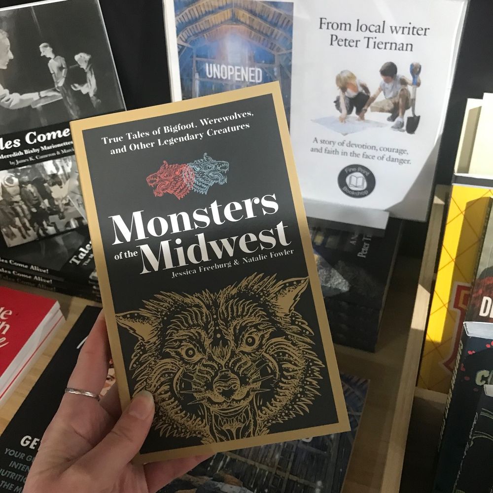 A picture of a white hand holding a book in front of a pile of other books. The book the hand is holding is Monsters of the Midwest. There's a line drawing of an unidentifiable monster on the cover.