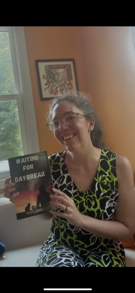 A white woman with frizzy brown hair wearing a green, white, and black dress holds a book up to the camera. The cover is visible, and it reads Waiting for Daybreak in white font. There is a silhouette of a cat and a woman on the cover, along with some blood splatter.