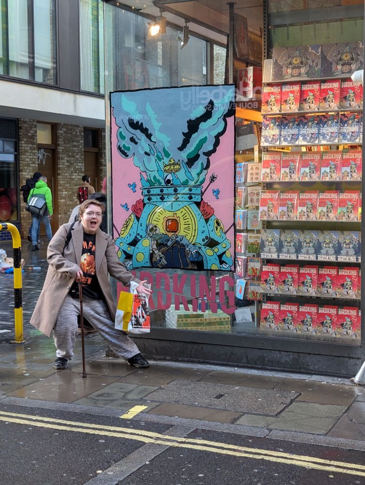 A man stands in front of a glass shop window. The shop window has a large painted mural in pink and blue. The street is rainy. The man is wearing a long brown coat, holding a walking stick in one hand and bags in the other hand, and is pointing to the mural while smiling widely at the camera.