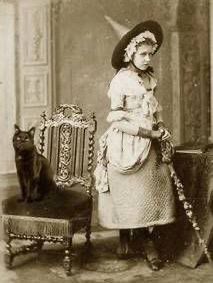 A turn of the century (1900) or earlier sepia photo of a girl in a pointed hat standing next to a fancy chair with a black cat sitting on it.