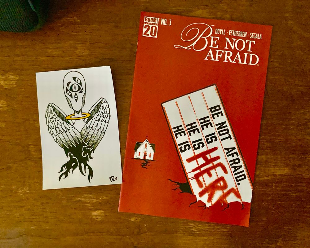 A copy of issue number three of the limited comic book series Be Not Afraid, written by Jude Ellison S. Doyle, illustrated by Lisandro Estherren, colored by Francesco Segala, and lettered by Simon Bowland. The cover is a uniform blood red, with a simple church house drawn in the background. In the foreground is a detached marquee, half stuck in the ground, that reads, 
