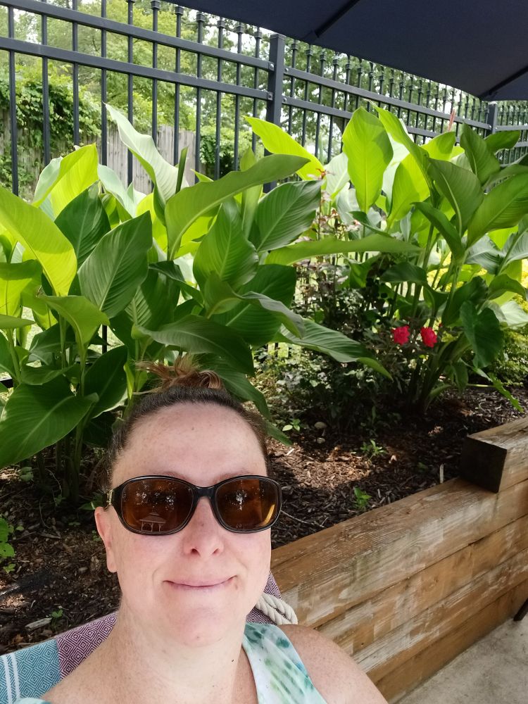 A girl in sunglasses sits under an umbrella next to a raised bed with canna lilies, rises and elephant ears. There is a black metal fence behind the plants. 