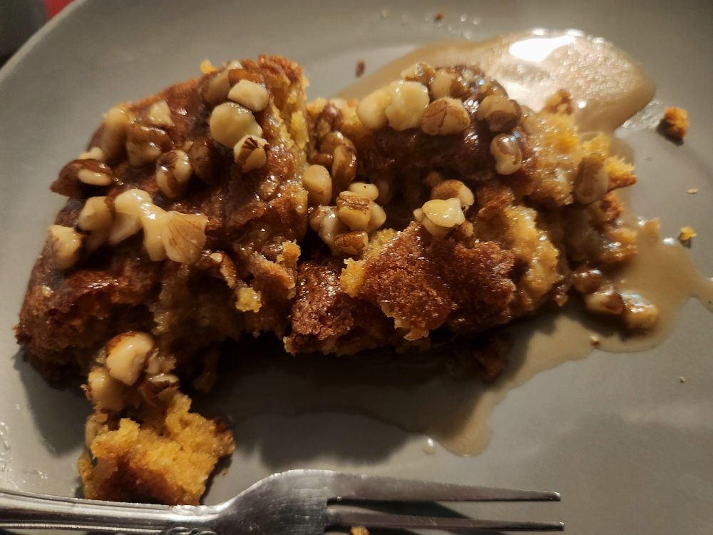Black walnut Blondie with maple sauce and black walnuts on top sitting on a plate next to a fork. 