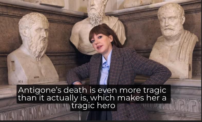 Philomena Cunk against a collection of classical statues. "Antigone's death is even more tragic than it actually is, which makes her a tragic hero"