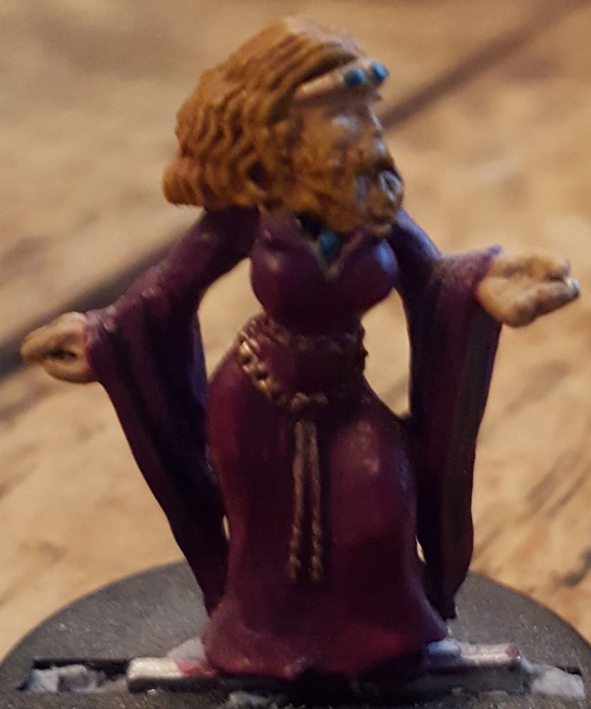 photo of a miniature figure of Tressa Quicksand: a woman with pronounced curves wearing a red-violet velvet dress with long draped sleeves. She's got strawberry blonde hair, mustache, and beard that is wavy with decorative braids. She has gold and teal jewelry