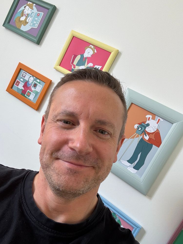 Paul, smiling in the Pooka office, in front of a series of photo frames containing the Pooka rabbit mascot doing things like taking photos, and using digital tools