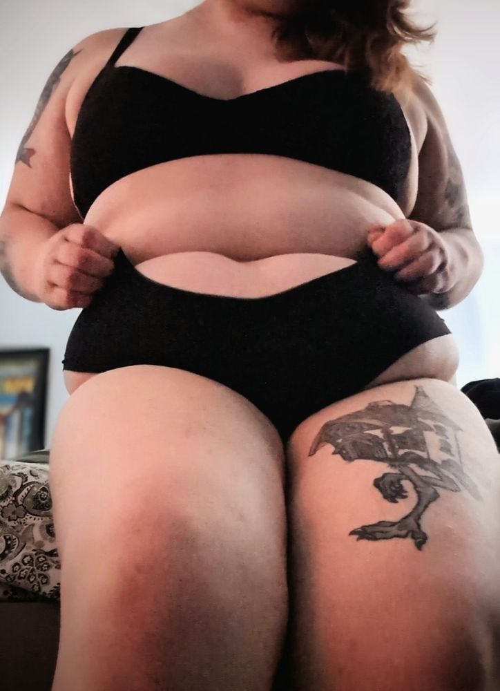 Fia sitting at the edge of a bed wearing black underwear 