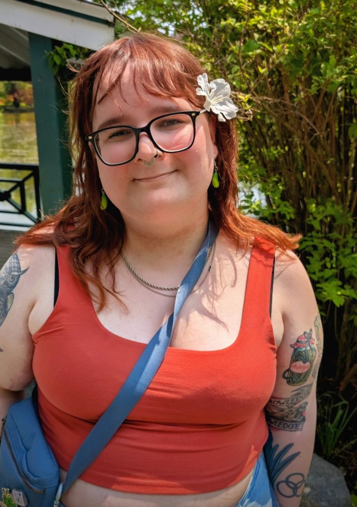 Fia outdoors in a red tank top. She has pickle earrings and a white flower in her hair 