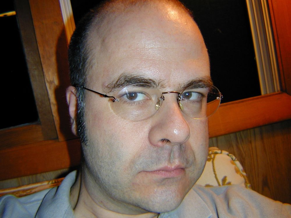 I am facing the camera, you see my head neck and collar. Im sitting in front of a dark window. I’m bald and clean shaven, except for sideburns, wearing rimless glasses. I’m wearing what looks like a gray polo shirt. I have a deep philtrum. 