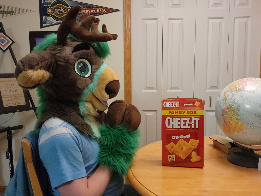 A green, grey fursuiter sitting at a table.  He has his hooves together in anticipation of eating some of the Cheez-its on the table in front of him. 