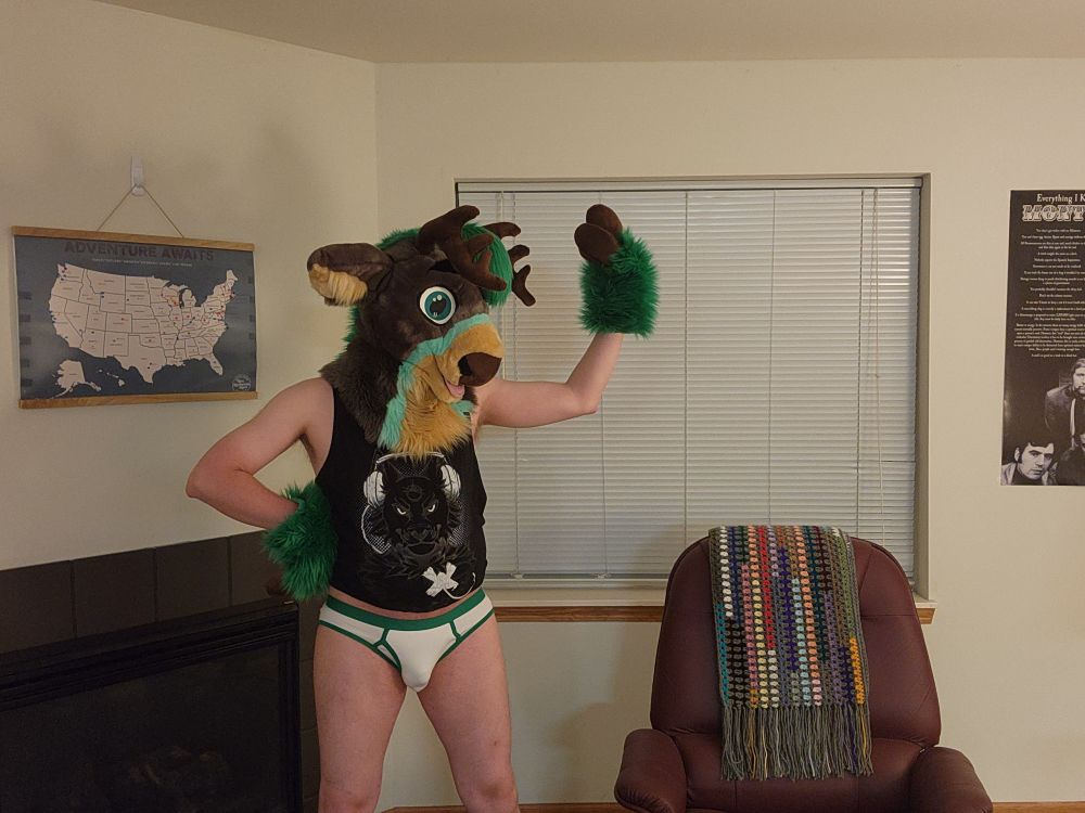 Green / grey partial fursuiter with only a fursuit head and hand paws wearing a black tank top with white briefs with green trim.  One hoof-hand is on his hip.  The other is raised as he tries to flex his muscles.  