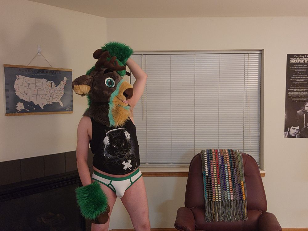 Green / grey partial fursuiter with only a fursuit head and hand paws wearing a black tank top with white briefs with green trim.  One hoof-hand is raised high over his head.  He appears to be sniffing his armpit.  The other hand which was on his hip has now slid down to hang loosely by his side. 