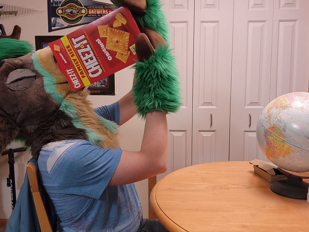A green, grey fursuiter sitting at a table.  In a moment of realization, he picks the box up, sticks his nose in, and tilts the box up!  That is how he can get to his precious Cheez-its!  