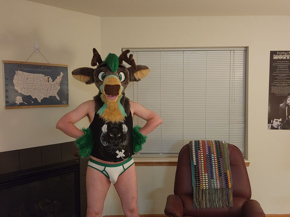 Green / grey partial fursuiter with only a fursuit head and hand paws wearing a black tank top with white briefs with green trim.  His hoof-hands are on his hips.  He is standing and looking at the camera.  