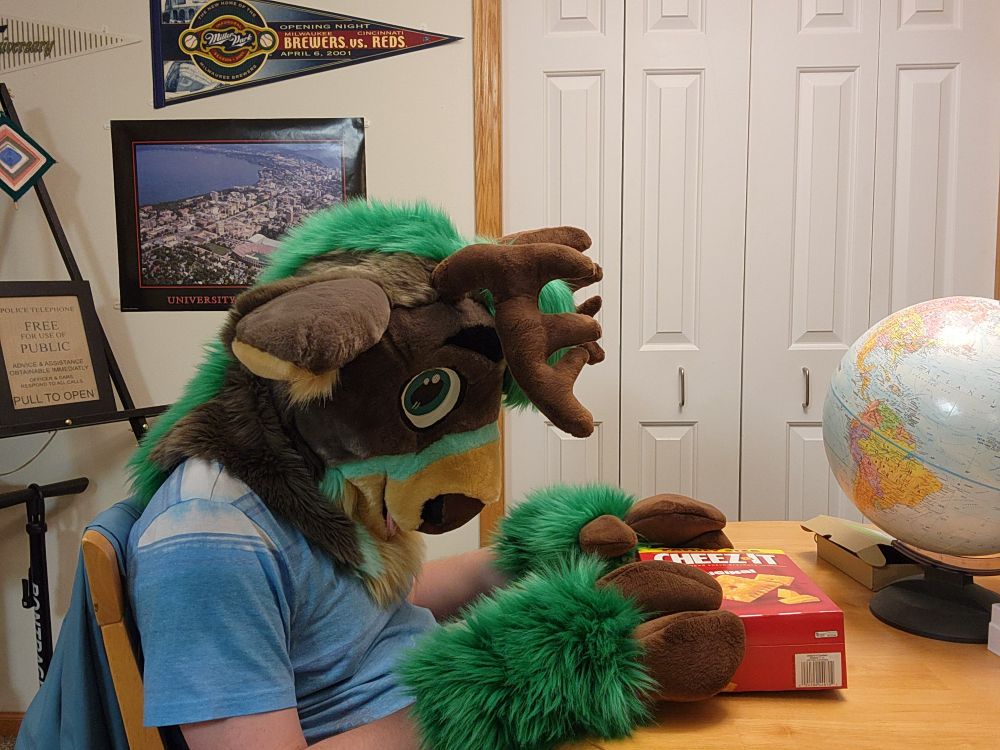 A green, grey fursuiter sitting at a table.  He set the box down on the table and is looking down, defeated.  How will he get the Cheez-its out of the box?  