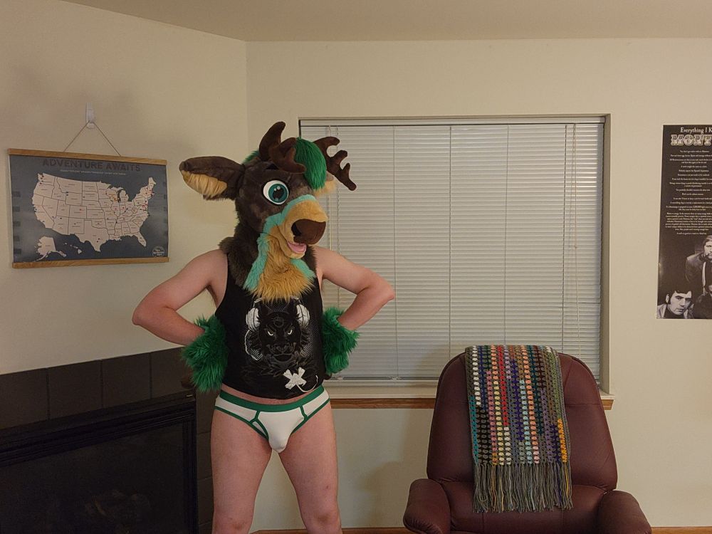 Green / grey partial fursuiter with only a fursuit head and hand paws wearing a black tank top with white briefs with green trim.  His hoof-hands are on his hips.  He is standing and looking off to the side.  