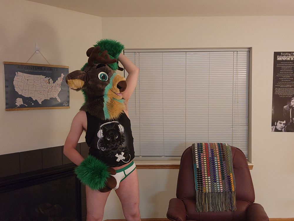 Green / grey partial fursuiter with only a fursuit head and hand paws wearing a black tank top with white briefs with green trim.  One hoof-hand is raised high over his head.  He appears to be sniffing his armpit.  The other hand which was on his hip has now moved to rest on his undie bulge.  