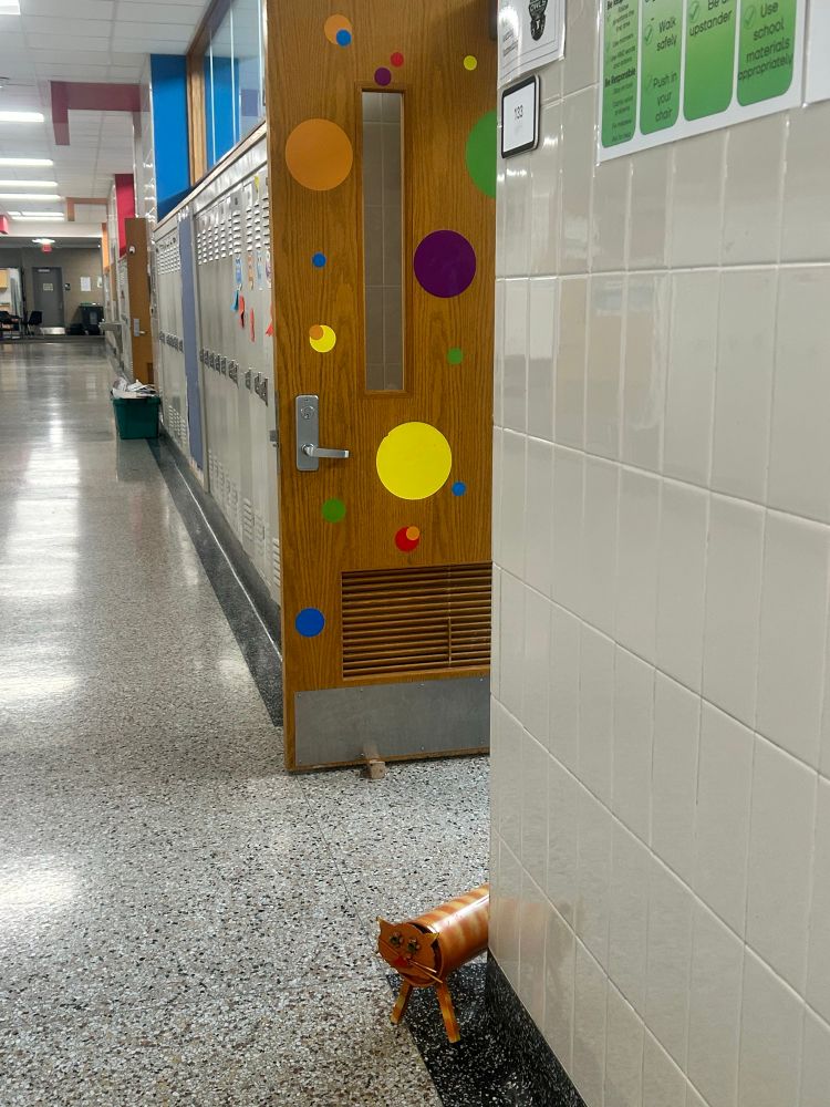 An elementary school hallways. A doorway with a propped open door that has colorful dots on it has a metal orange cat peeking around the corner