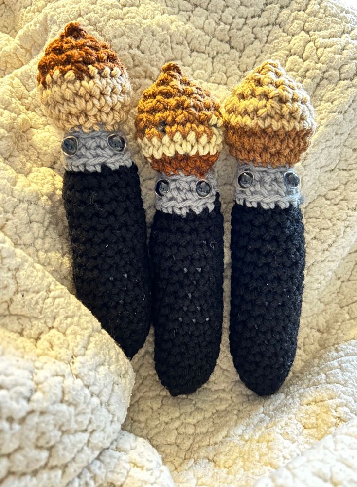 A trio of crochet paintbrushes on a fleece blanket. They have a black handle, gray with eyes then bristles of various brown/tans