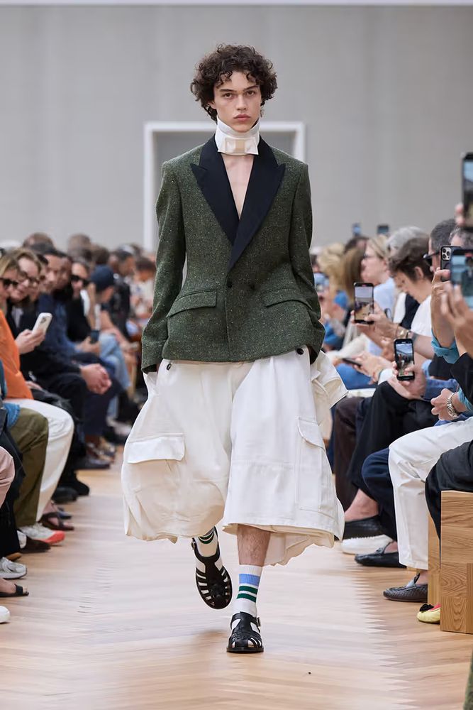 photo of a model from the dior menswear ss26 show. the model is wearing a white neck scarf, a green jacket, white baggy pants, white socks with blue stripes and black sandals.