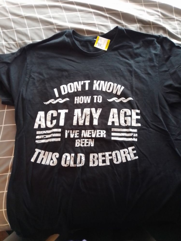 A black T-shirt laying on a grey gridded bedsheet. The shirt reads "I don't know how to act my age, I've never been this old before." 