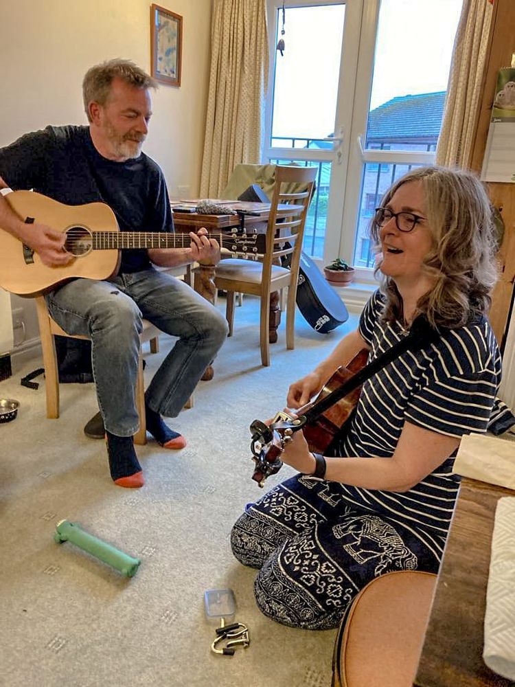 A man and woman are playing guitars in a bright sunlit room, the man is playing a mini travel guitar and sitting on a chair, the woman is kneeling on a carpet whilst singing.