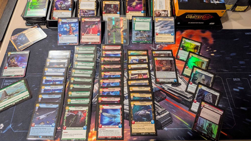 A tabletop with a Star Wars Unlimited themed playmat, numerous trading cards in piles, and a box of "Jump to Lightspeed" booster packs.
