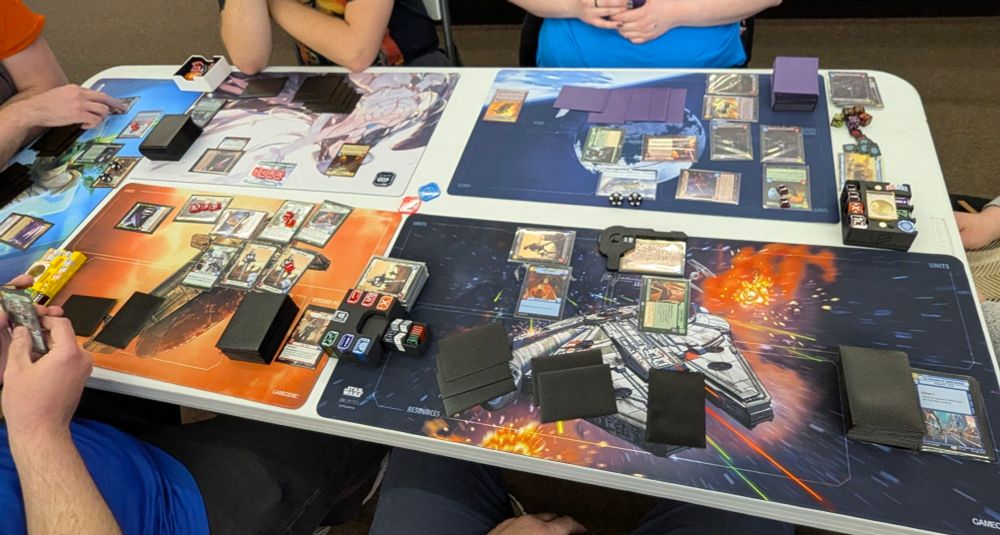 A high-angle, slightly distorted shot of a rectangular table from above, with five players playing the Twin Suns format of Star Wars Unlimited, a TCG game. The table is covered with five colorful playmats, each with a different design, and various cards are spread across them. Players' hands and forearms are visible around the edges of the table, suggesting they are in the midst of a game.