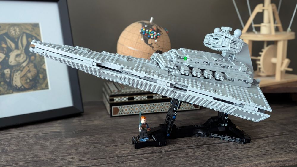 A fully assembled grey Lego Star Destroyer model on a black display stand, with a mini-figure at the base, set on a wooden surface.