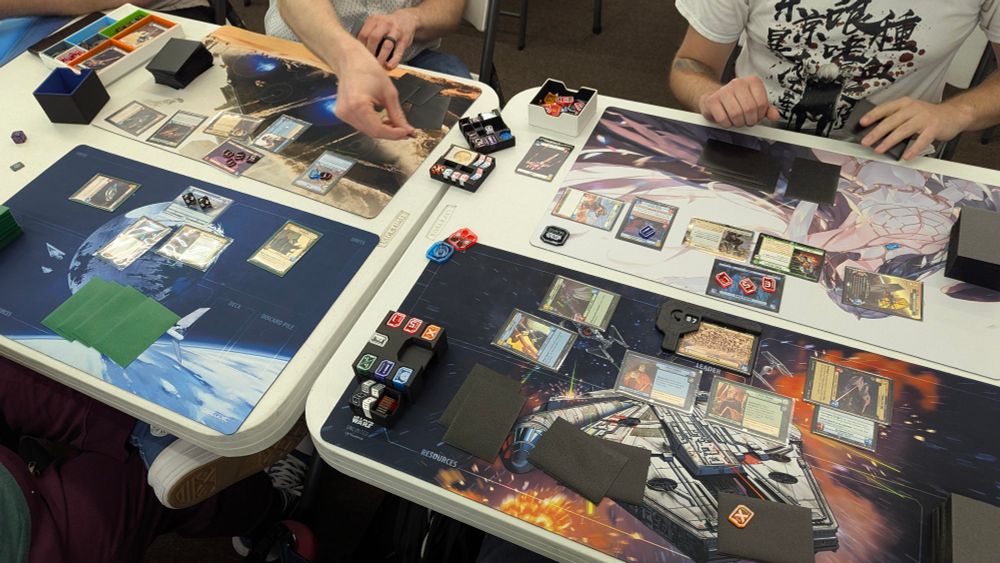 This image shows a tabletop scene with four players engaged in playing Twin Suns, a gameplay version of the collectable card game Star Wars Unlimited. 