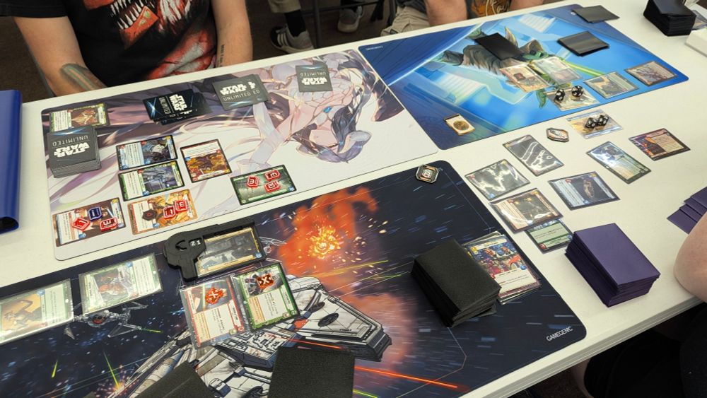 A top-down, slightly angled shot of a white table where three people are playing a card game. The table is covered with three playmats, each with different artwork. The playmat on the left has a dark background and features a Star Wars Unlimited logo, along with various cards laid out in rows. The center playmat has a light background with an anime-style female character, and also has Star Wars Unlimited cards arranged on it. The playmat on the right has a fiery design and more cards, including some dice and tokens.  Each player has a stack of cards, some face up and some face down, indicating an active game in progress.