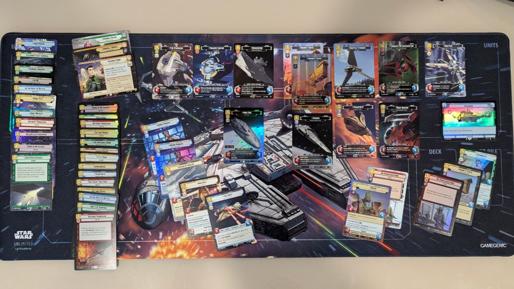 A top-down shot of a Star Wars Unlimited card game layout on a dark gray playmat. The playmat features a detailed illustration of a large, gray spaceship with various smaller ships around it, set against a backdrop of space.  Scattered across the playmat are numerous Star Wars Unlimited cards, arranged in distinct piles and rows.  On the left side, a vertical stack of cards is partially visible, with the "Star Wars Unlimited" logo at the bottom.  Moving right, a row of face-up cards is displayed, featuring various characters and vehicles from the Star Wars universe.  Below this row, another set of cards is spread out, with some overlapping.  On the right side of the playmat, two areas are labeled "UNITS" and "DECK," each with a stack of cards beside it.  The "GAMEGENIC" logo is visible in the bottom right corner of the playmat.