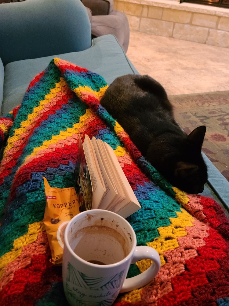 Legs on an aqua couch, covered with a bright rainbow blanket, black cat, mug of cocoa, paperback book, and a bag of chocolate coffee beans.
