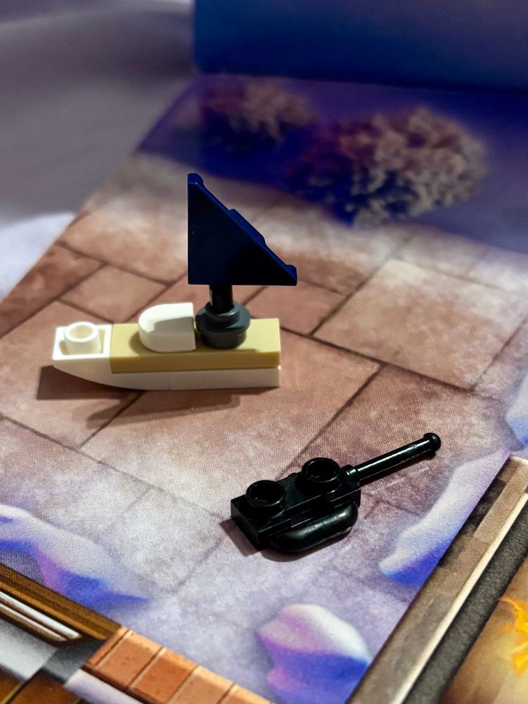 A tan and white Lego sailboat with navy sail. A black Lego remote control sits next to it. 