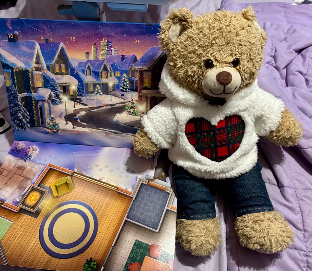 A teddy bear sitting with a Lego advent calendar. It has a ski chalet village scene where the doors are and an attached display board with a ski lounge scene. 
