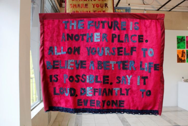 A red banner with black cut out letters reading 'The future is another place. Allow yourself to believe a better life is possible. Say it loud, defiantly to everyone'