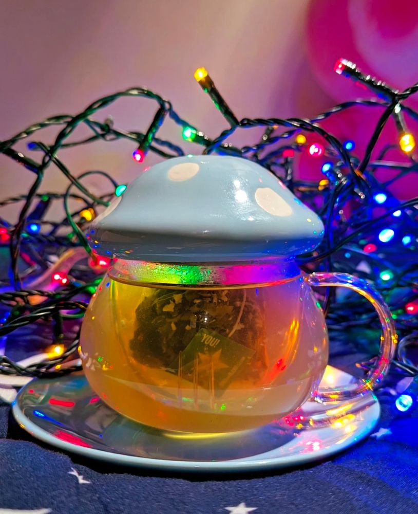A tea glass shaped like a toadstool with a ceramic lid in baby blue with white spots on a baby blue ceramic saucer. A bundle of multicoloured (blue, red, green, yellow) led xmas lights are in the background. 