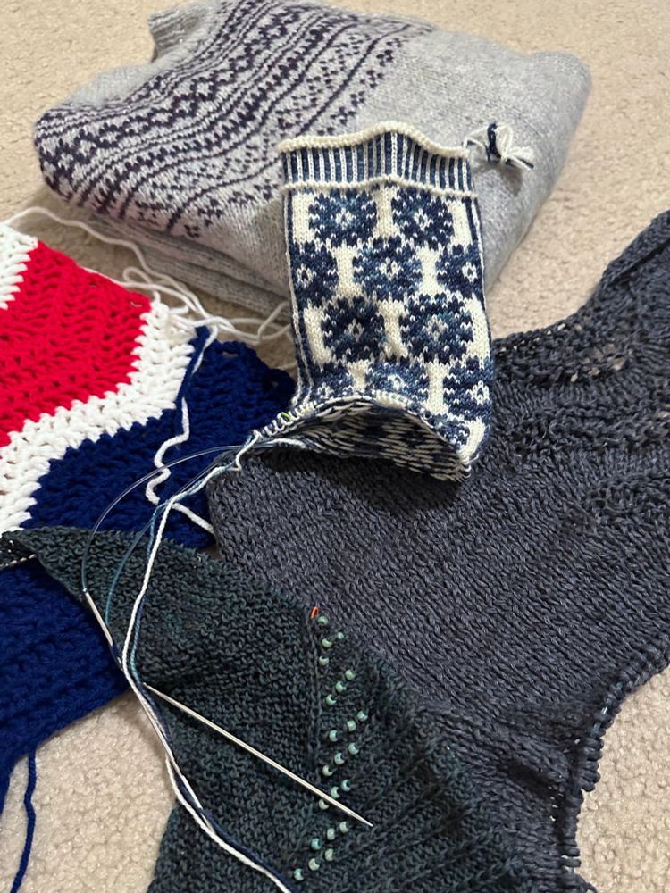 Photo of several partially completed knit or crochet projects. A pale gray-blue cardigan with a dark purple stranded yoke, folded and all done except finishing. A stranded colorwork sock in white with dark blue flowers or starbursts. A dark gray stockinette panel with an old shale lace pattern down the middle. A petrol green asymmetric triangular shawl with garter on the left and lines of twisted knits ending in leaves at the shawl edge, with turquoise beads on. A blue, red, and white striped ripple crochet afghan.
