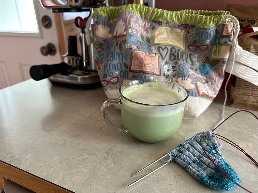 A half-knit sock (toe-up magic loop) with its project bag sitting on a kitchen counter with a large matcha green tea latte in a glass mug. An espresso machine is in the background. 