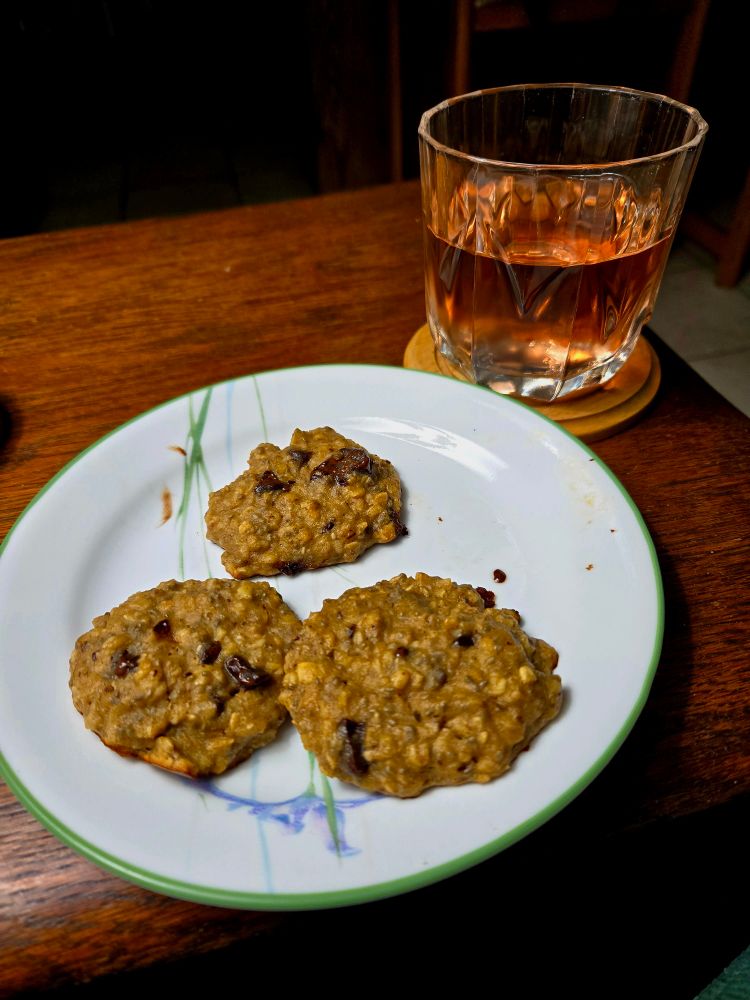 Photograph of banana, oatmeal, chia & chocolate chip cookies on a plate, plus a glass of rosé wine, all on an antique wooden table. 