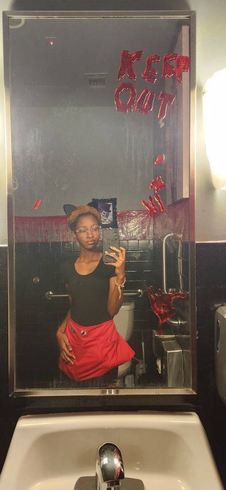a black feminine person wearing a black t shirt and a red half apron. Their hair is short and bleached and they are wearing cat ears. They are taking a mirror selfie in the bathroom and on the mirror is Halloween decorations that says "KEEP OUT" and has fake bloody handprints.