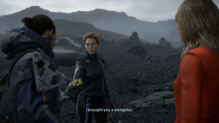 A screenshot from the end of Death Stranding. Three people are standing on a volcanic beach. A woman, Amelie, stands to the right. A man to the left, Sam, is accepting a golden mask from a woman in the centre, Fragile, who is saying "I brought you a metaphor".
