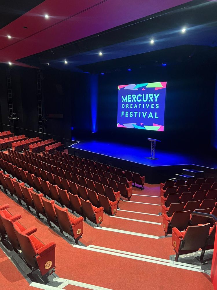 A picture of the seats in a theatre and a stage with a screen at the back displaying the text “Mercury Creatives Festival”