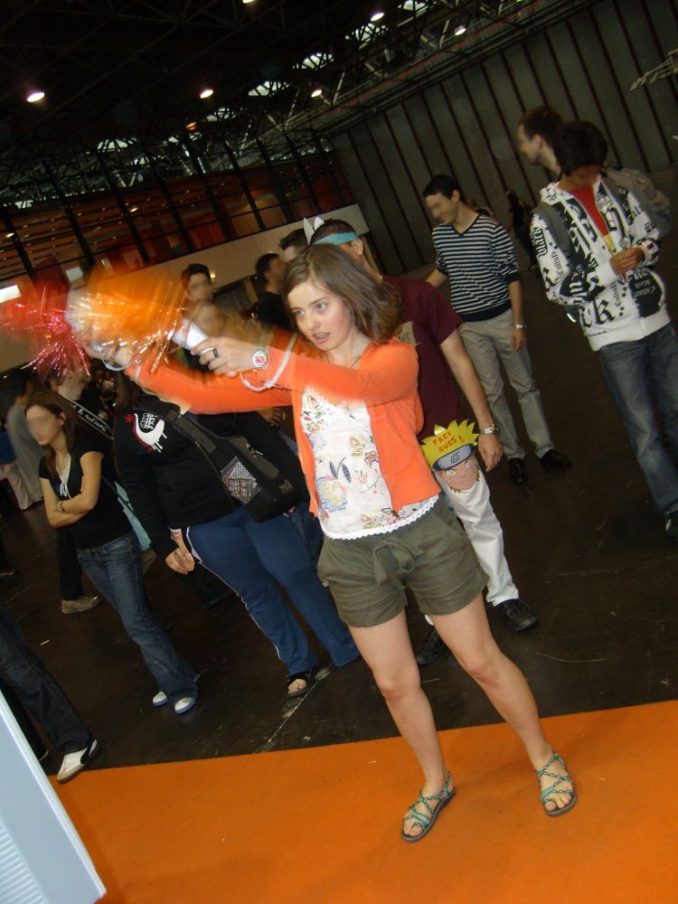 Lucie, 17 ans, agite des pompoms accrochés à des manettes de Wii. Son gilet TRÈS ORANGE est assorti à la moquette TRÈS ORANGE de l'espace jeux vidéo de Japan Expo.