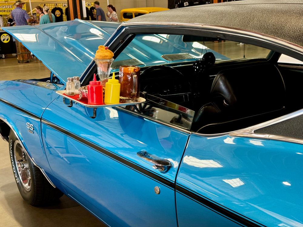 A blue car in a car show, with the driver’s side window rolled down to accept a Drive In food tray. Root beer, a float, and condiments on the tray. 