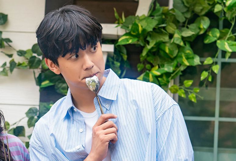 Shin Hyun Seung from The Potato Lab, wearing a blue shirt and with a spoon in his mouth, looking off to the right in front of a window with ivy growing across it. 