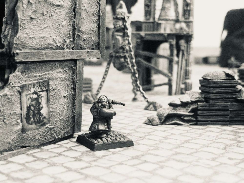 a photograph of a medieval gaming board; a dwarven crossbowman aims around a the corner of a dilapidated building